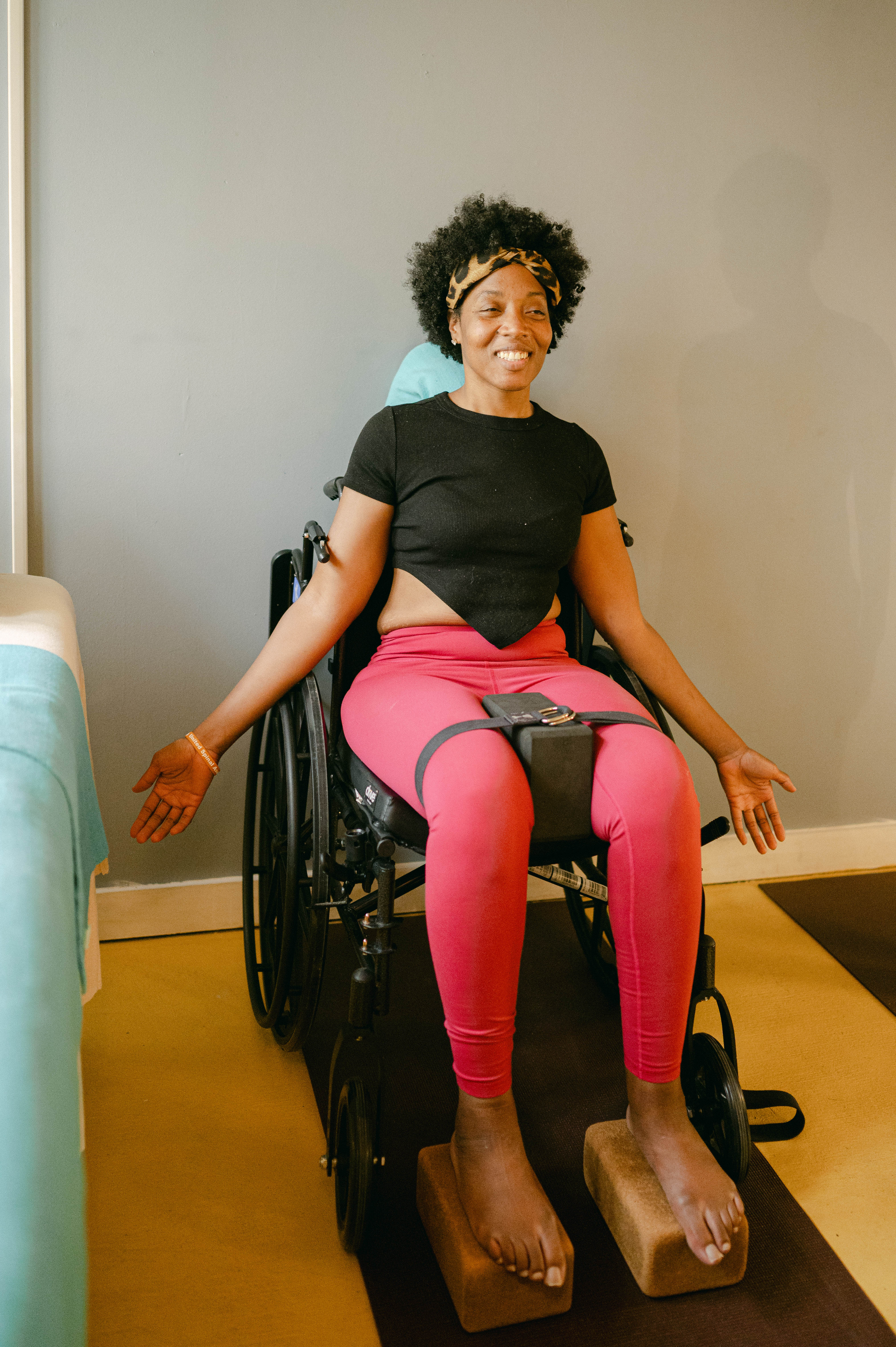 Ashley, a black woman with MS, is sitting in a black wheelchair. She's wearing a black crop top, pink leggings, and a cheetah print headband around her dark curly hair. She has a yoga block strapped between her thighs, two cork blocks under her feet, and she is positioned in mountain pose with her arms reached down by her sides. 