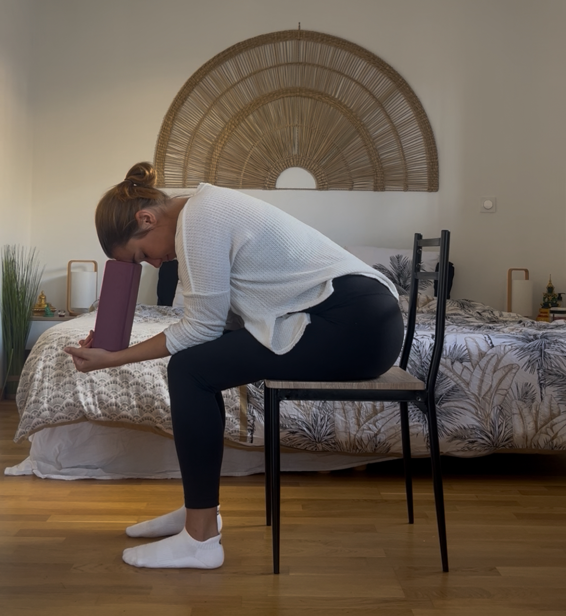 Caroline is seated in a chair doing a version of child's pose. She's wearing a white long sleeve top, black leggings, and white socks. She's leaning forward with her forearms rested on her thighs holding a yoga block that is supporting her forehead. In her opinion it's one of the best chair poses out there. 